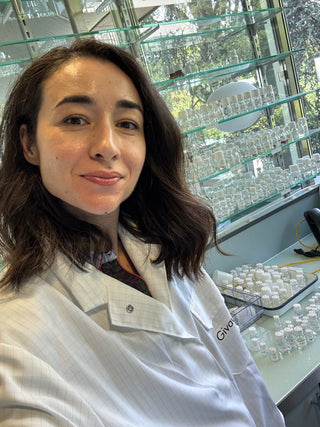 Founder in a lab coat standing in a laboratory setting with shelves of glassware and fragrances