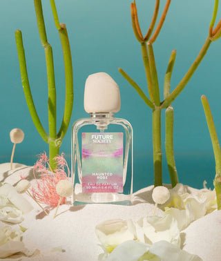 Perfume bottle labeled "Haunted Rose" on sand surrounded by flower petals and aquatic plants on blue background