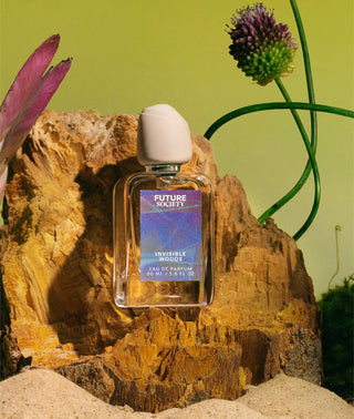Perfume bottle labeled "Invisible Woods" on jagged rock with sand in foreground surrounded by exotic flowers