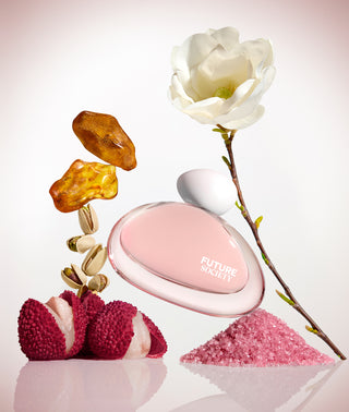 A perfume bottle with a pink oval body and clear cap, labeled 'FUTURE SOCIETY', styled with a white magnolia flower, amber stones, pink crystals, and lychee halves on a clean white background.