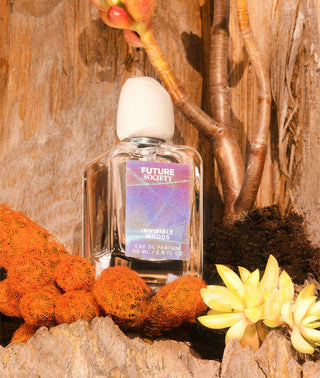 Perfume bottle labelled "Invisible woods" against rocky backdrop with exotic flowers in foreground